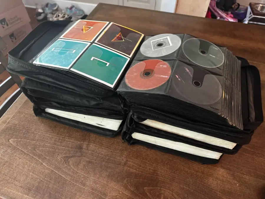 A black CD binder sits open on a wooden table, displaying several CDs and album art sleeves. Boxes and shoes are visible in the background.