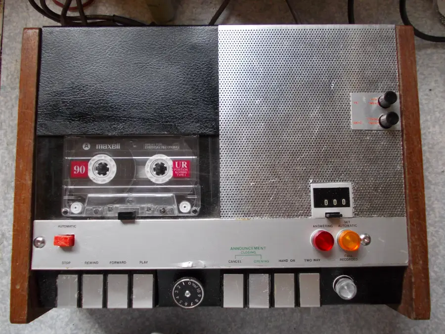 A vintage tape recorder with a cassette tape inside, various buttons, switches, indicator lights, a dial, and a small analog display on a metallic and wood-paneled surface.