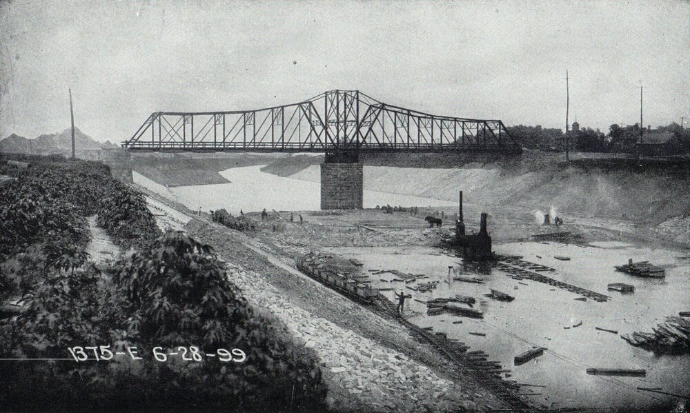 Black-and-white photo of a metal truss bridge over a river, with people and logs near the water’s edge. Date “6-28-99” is written at the bottom, indicating June 28, 1899.