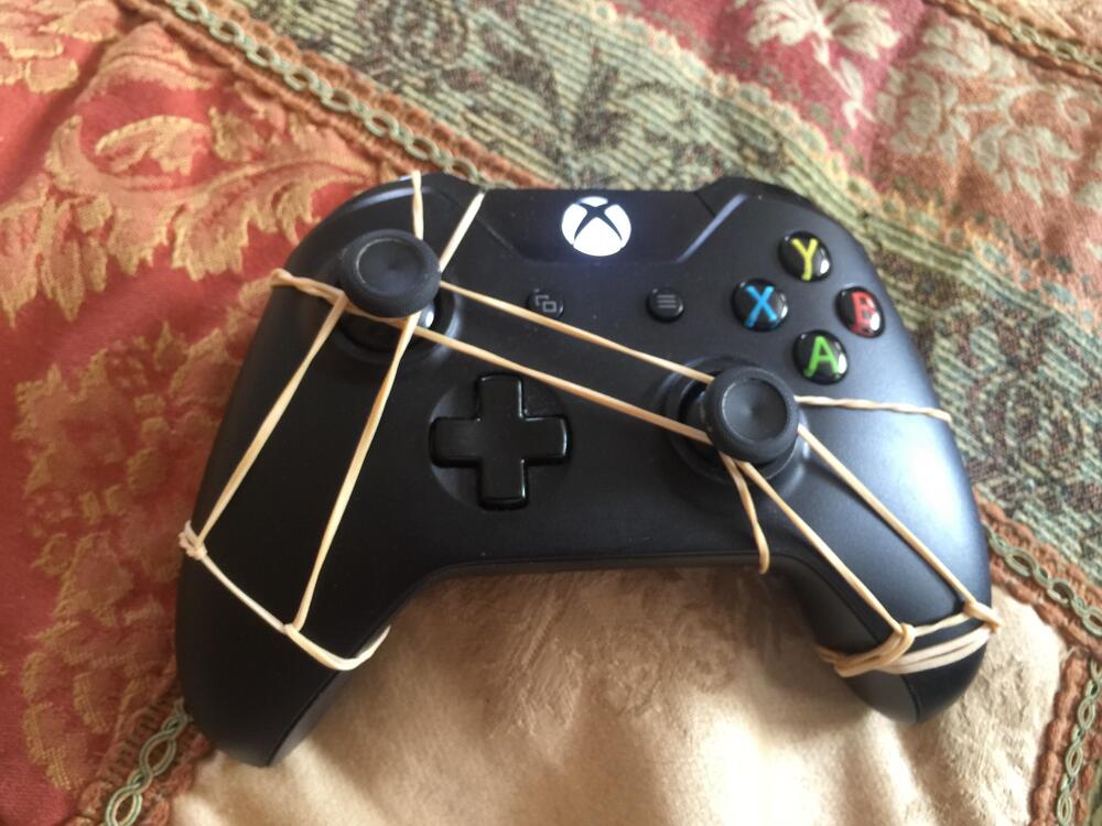 A black Xbox controller with several rubber bands wrapped around it, placed on a patterned fabric surface and a tan cloth.