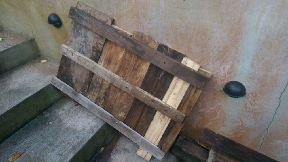 A wooden pallet with various shades of brown planks leans against a textured concrete wall near outdoor steps. Two round black wall lights are mounted on the wall.