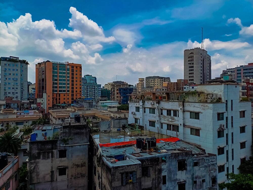 Cityscape of Dhaka under a bright blue sky with white clouds.