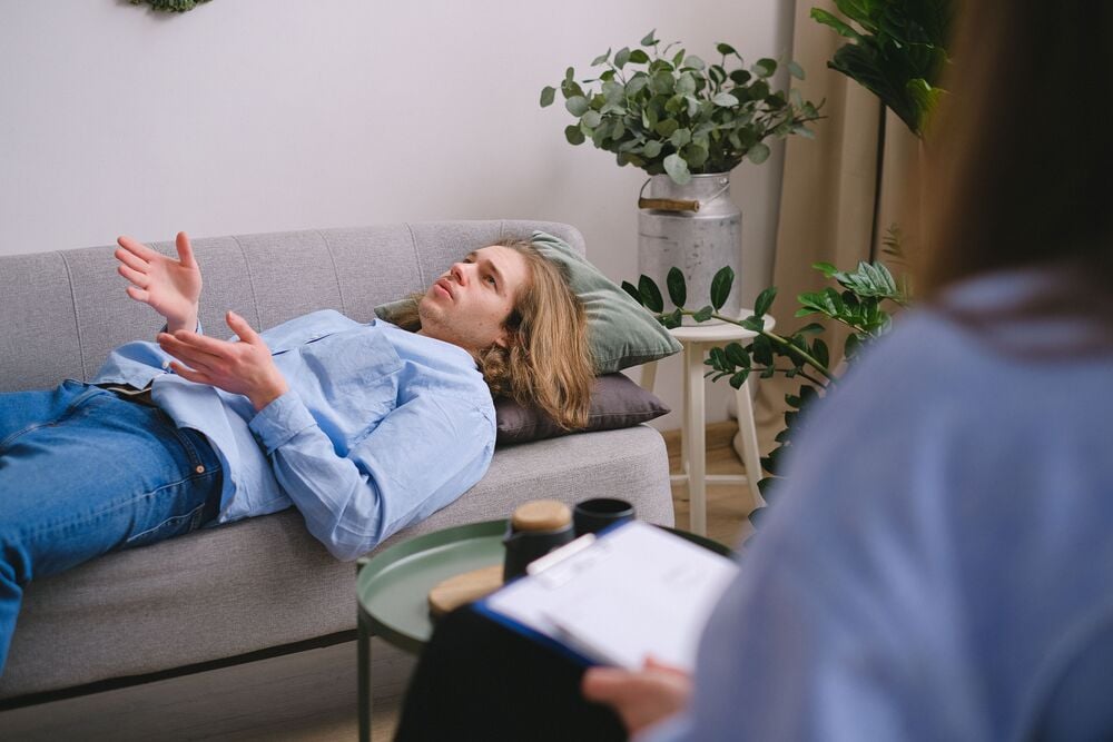 A person lies on a couch talking while another person takes notes on a clipboard.