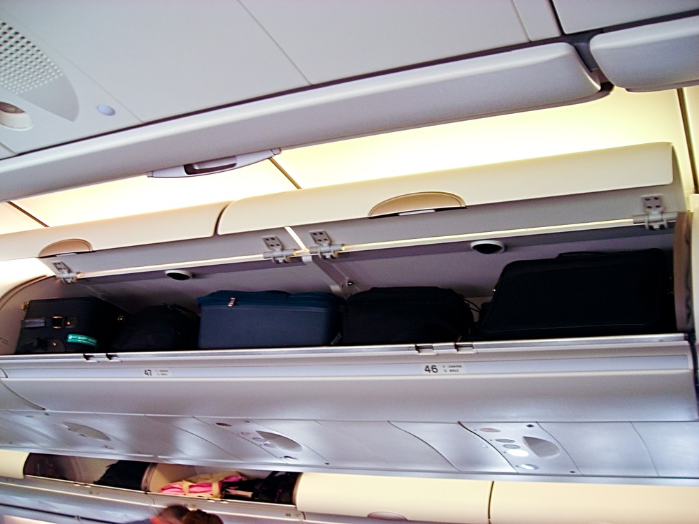 Open airplane overhead bin filled with several black suitcases and bags. The cabin ceiling and closed bins are visible below the open compartment.