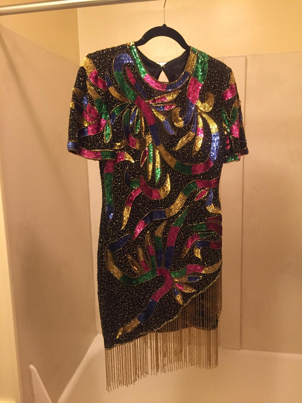 A short-sleeve black dress covered in colorful sequins with wavy patterns in gold, blue, green, and pink, hanging on a hanger. The dress has fringe detailing at the bottom.