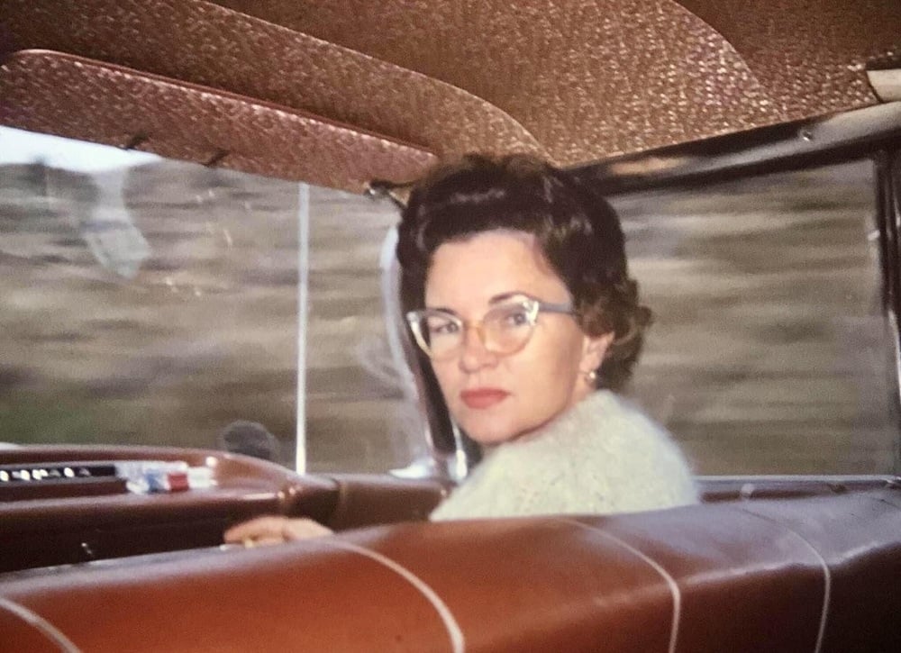 A woman with glasses and dark hair sits in the front seat of a vintage car, looking back over her shoulder. The car has brown leather seats and a blurred landscape is visible outside the window.