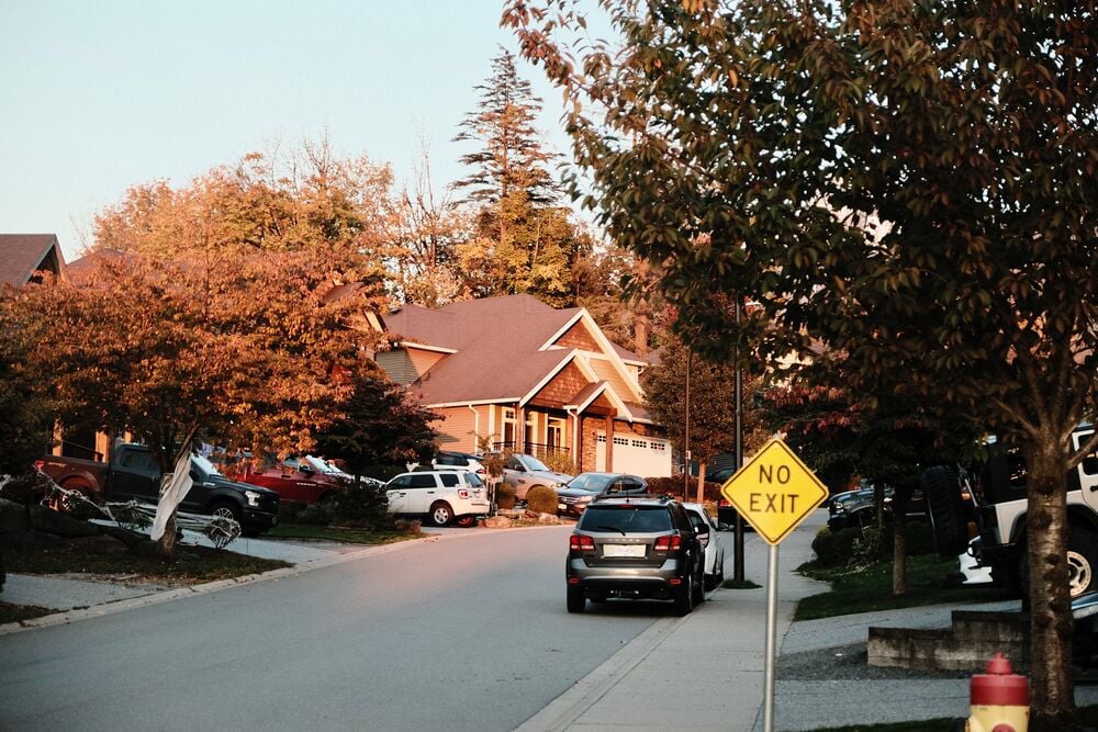 A suburban street with parked cars, autumn trees, a fire hydrant, and a yellow “No Exit” sign. Houses with porches are visible under a clear sky in the background.