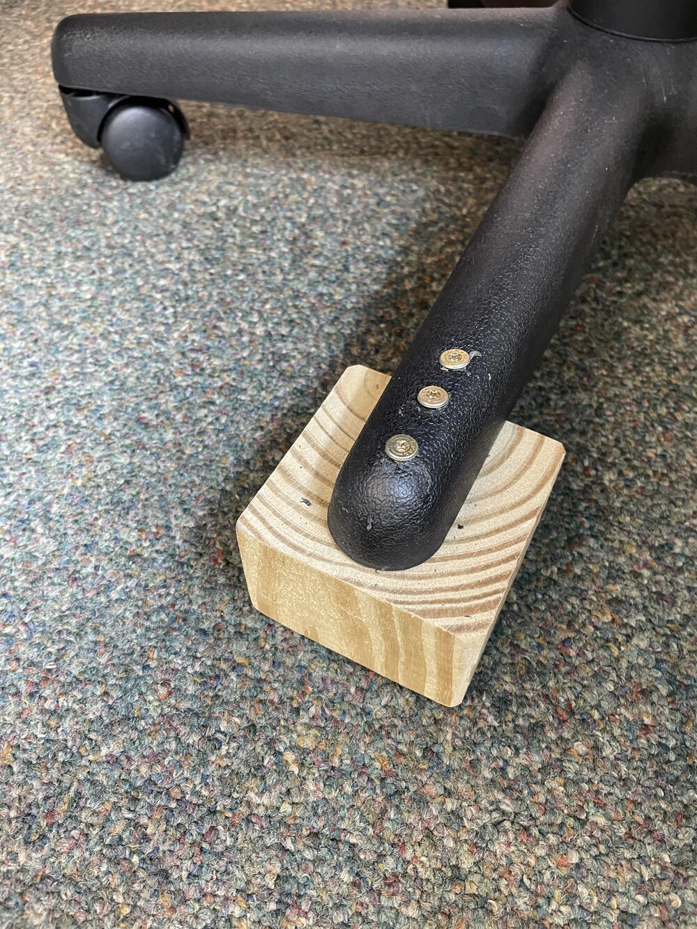 A black office chair leg is attached to a small wooden block using three screws, placed on a speckled carpeted floor.
