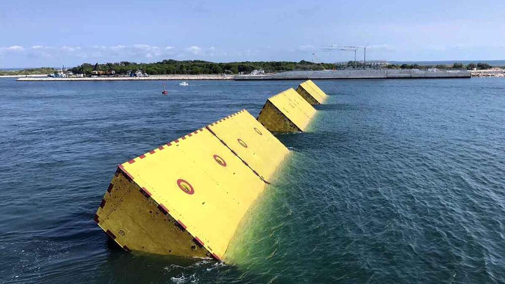 Large yellow floating barriers rise at an angle from the blue water near a shoreline, under a clear sky, with land and construction cranes visible in the distance.