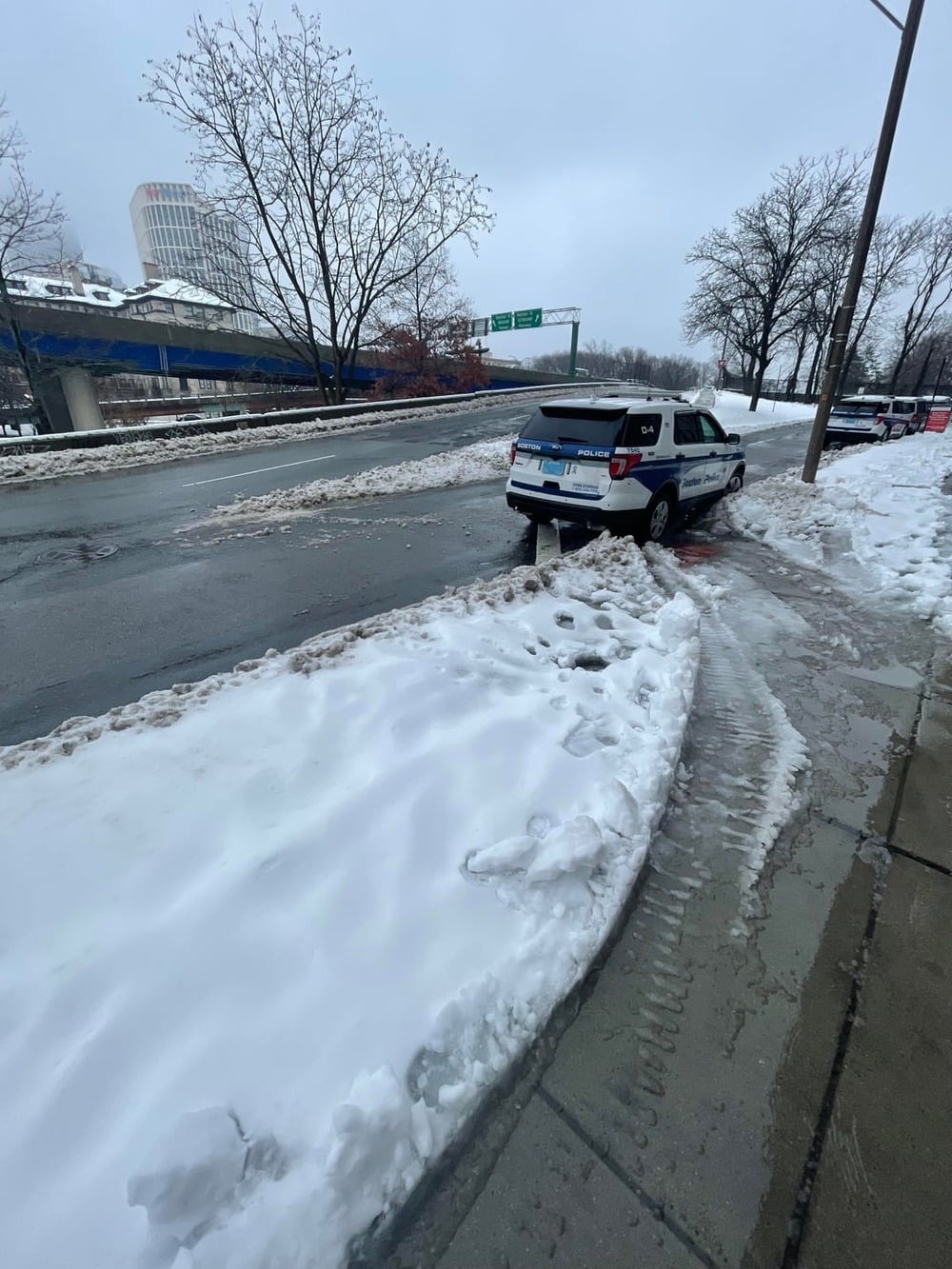 A police SUV is parked on a snowy curb near a wet road, with snow-covered sidewalks and bare trees visible under a gray sky.