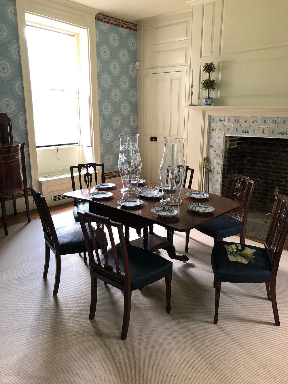 A formal dining room with a table set for six, blue wallpaper, a tiled fireplace, and large vases.