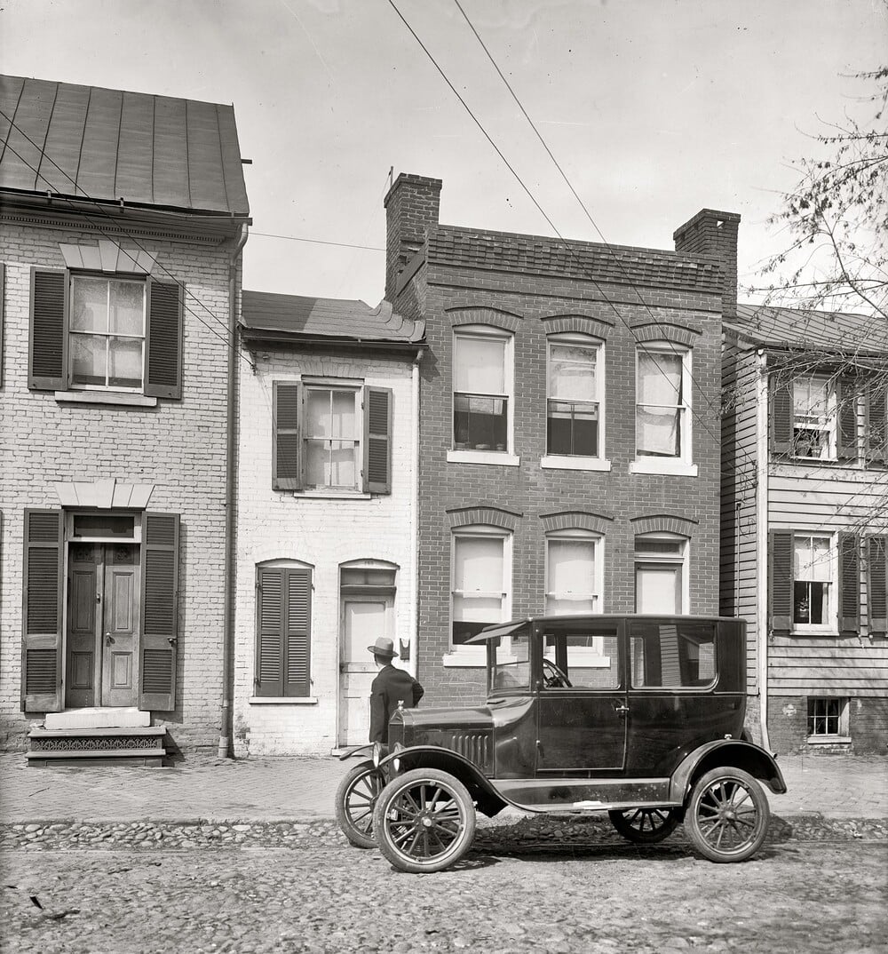A vintage car is parked on a cobblestone street in front of brick row houses, with a person standing next to the car and looking at the buildings. The scene appears to be from the early 20th century.