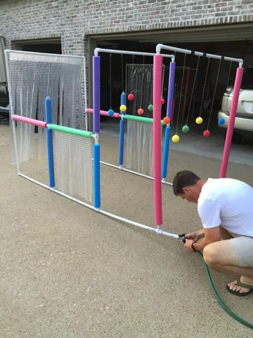 A man attaches a garden hose to a homemade PVC pipe water play structure with colorful foam and hanging balls, set on a driveway in front of a garage.