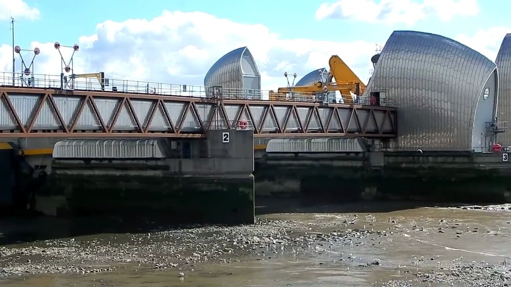 A large flood barrier with silver, curved gates spans a river during low tide, exposing the muddy riverbed. Equipment and vehicles are visible on the barrier under a partly cloudy sky.