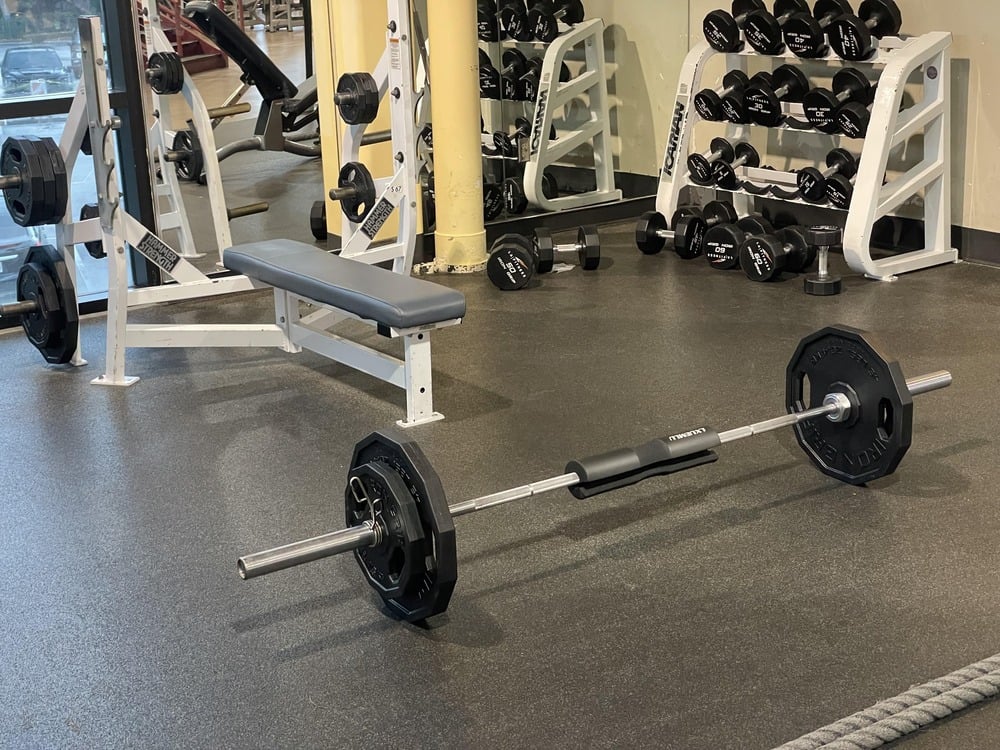 A gym with weight racks, a bench, and a barbell on the floor with padded grips. Dumbbells are organized on racks against the mirrored wall.