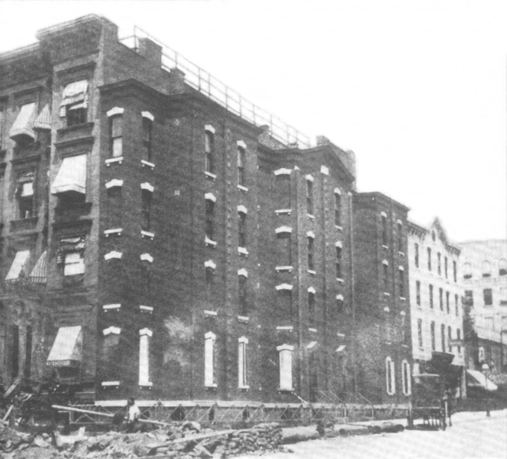 Black and white photo of old brick apartment buildings with broken windows and debris on the sidewalk. A horse-drawn carriage is visible on the street in front. The scene appears rundown and deserted.