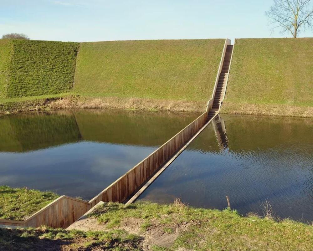 A narrow sunken wooden walkway cuts through a grassy embankment and water, resembling a “water bridge,” with stairs leading up the slope in the background under a blue sky.