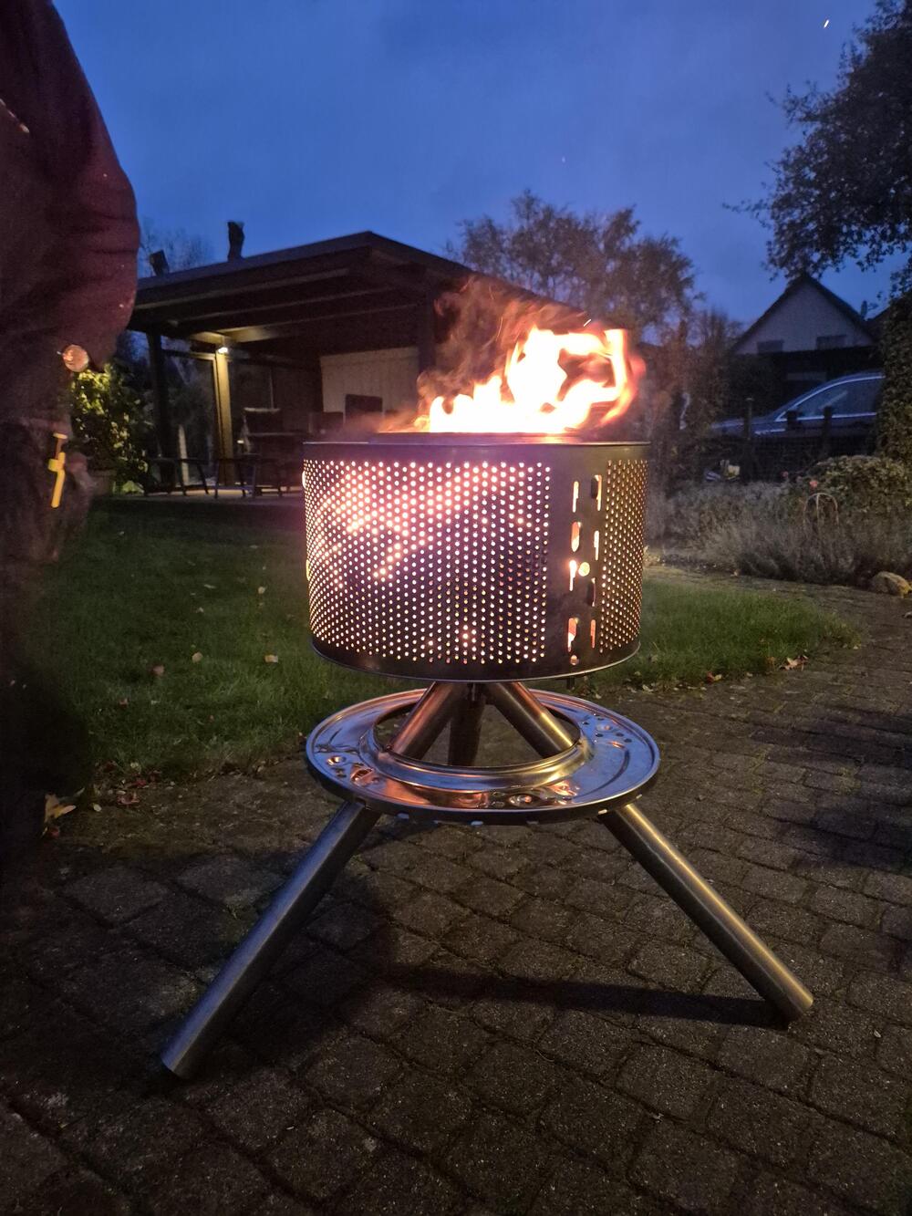 A metal drum, repurposed from a washing machine, is used as a fire pit. Flames rise from the drum, set on a stand outdoors in a backyard during dusk, with houses and trees in the background.