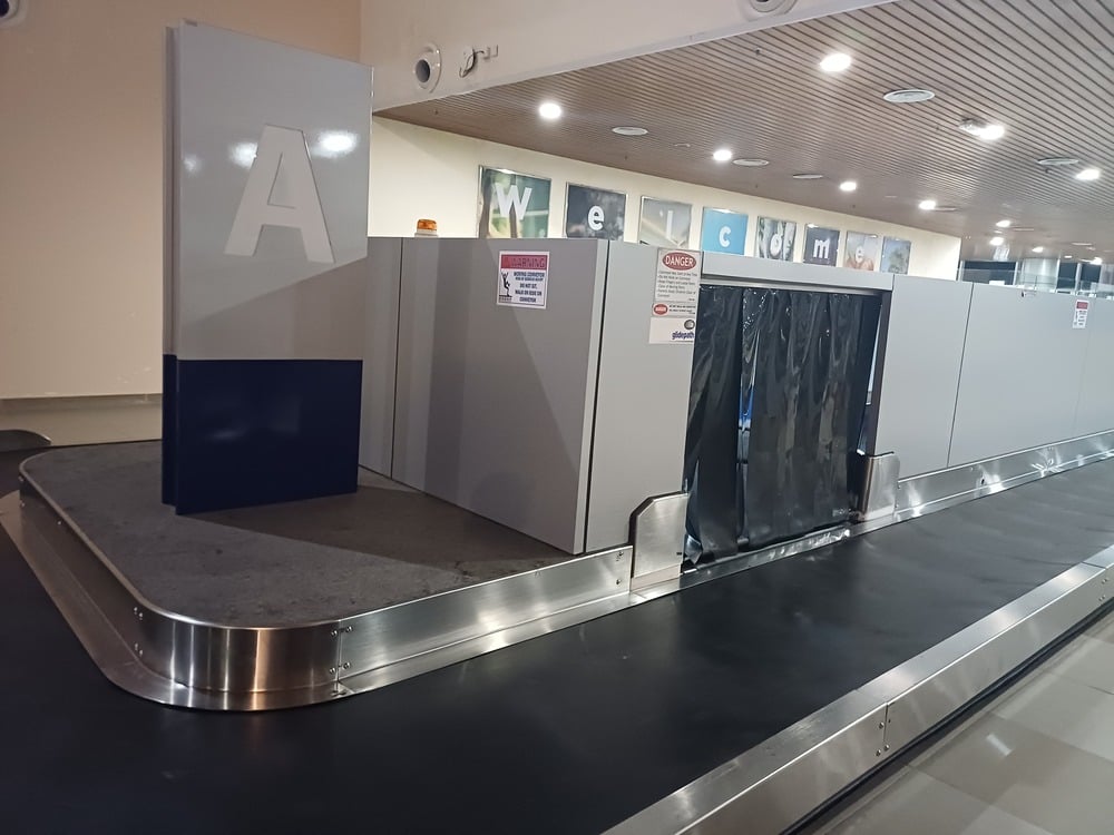 An empty airport baggage carousel, marked with a large letter “A,” stands still with no luggage on it. A welcome sign is visible in the background above the area.
