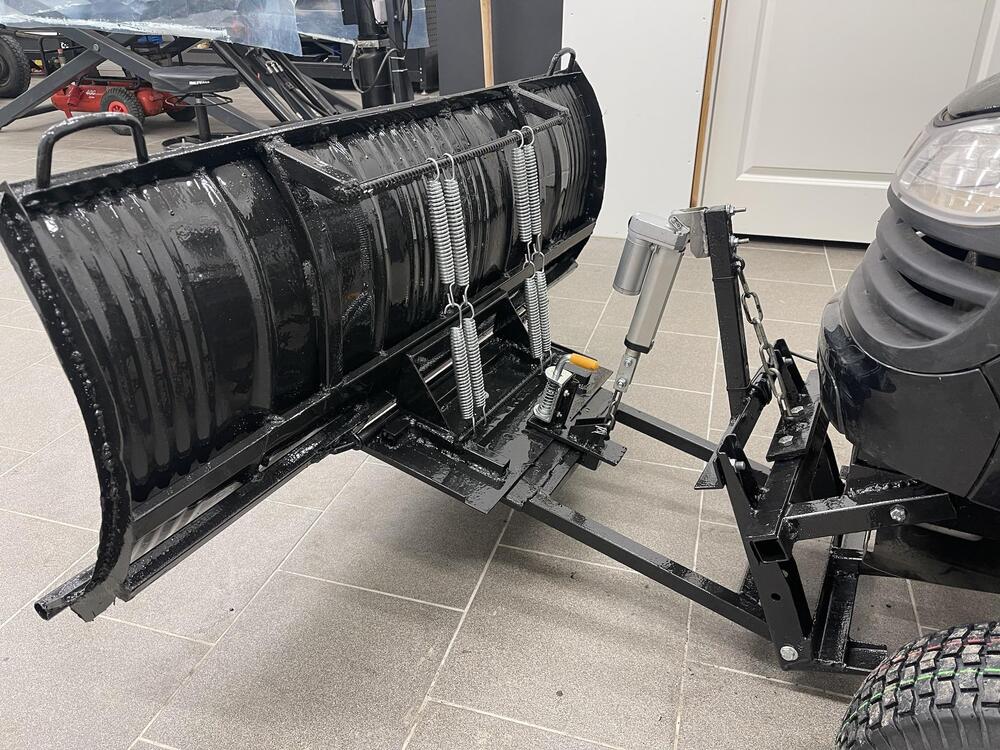A black metal snow plow attachment is mounted on the front of a small vehicle, shown indoors on a tiled floor. The plow has springs, brackets, and a hydraulic piston for angle adjustment.