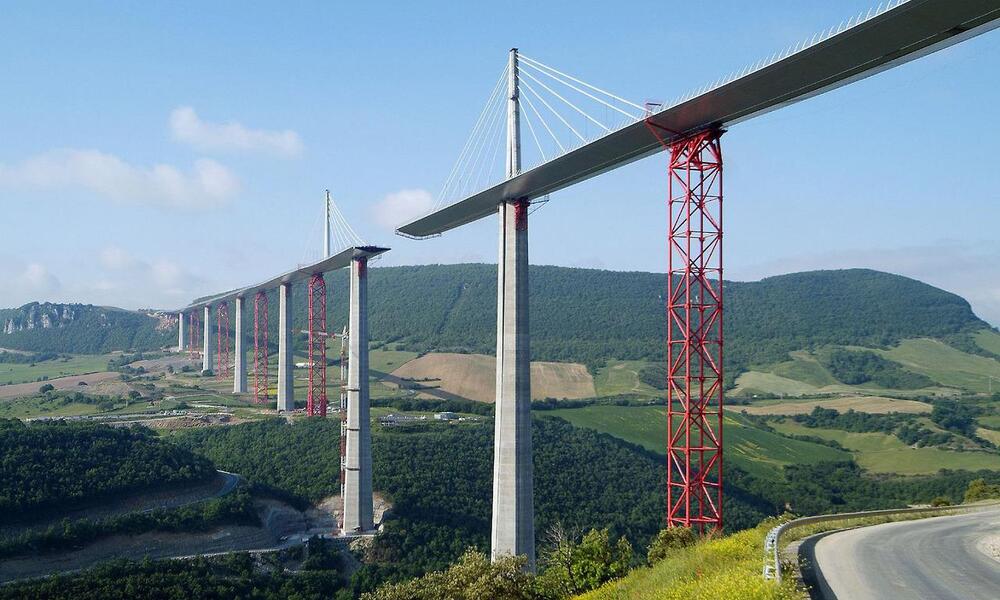 A long cable-stayed bridge with tall concrete pillars and red support structures spans a deep green valley, with hills and fields in the background under a blue sky.