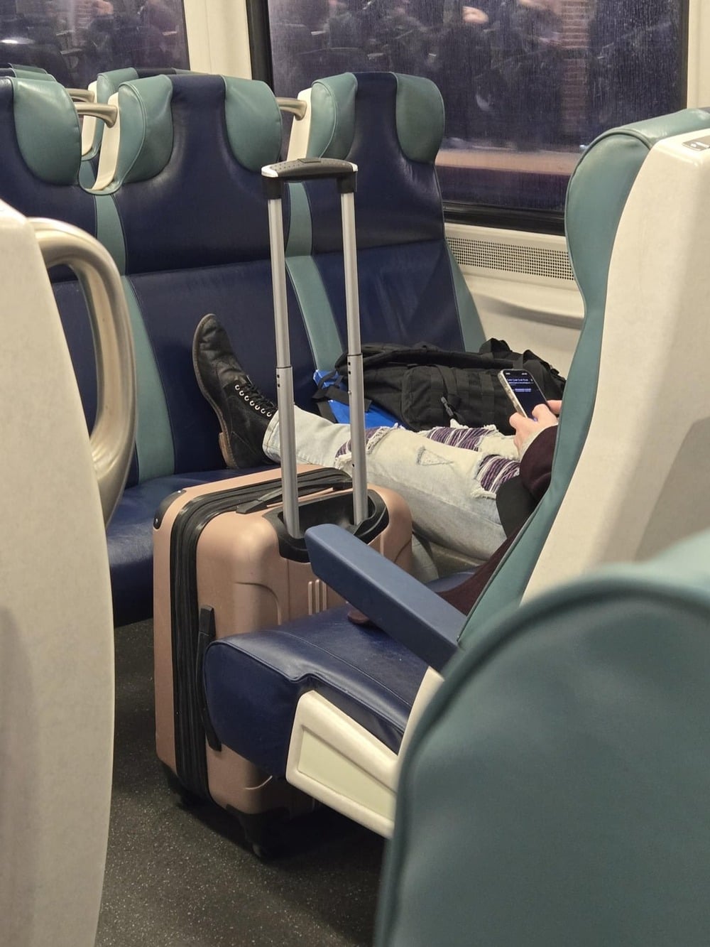 A person rests with legs stretched across train seats, boots on, suitcase in front, using a phone. The train is mostly empty, with blue and green seats and blurred scenery outside the window.
