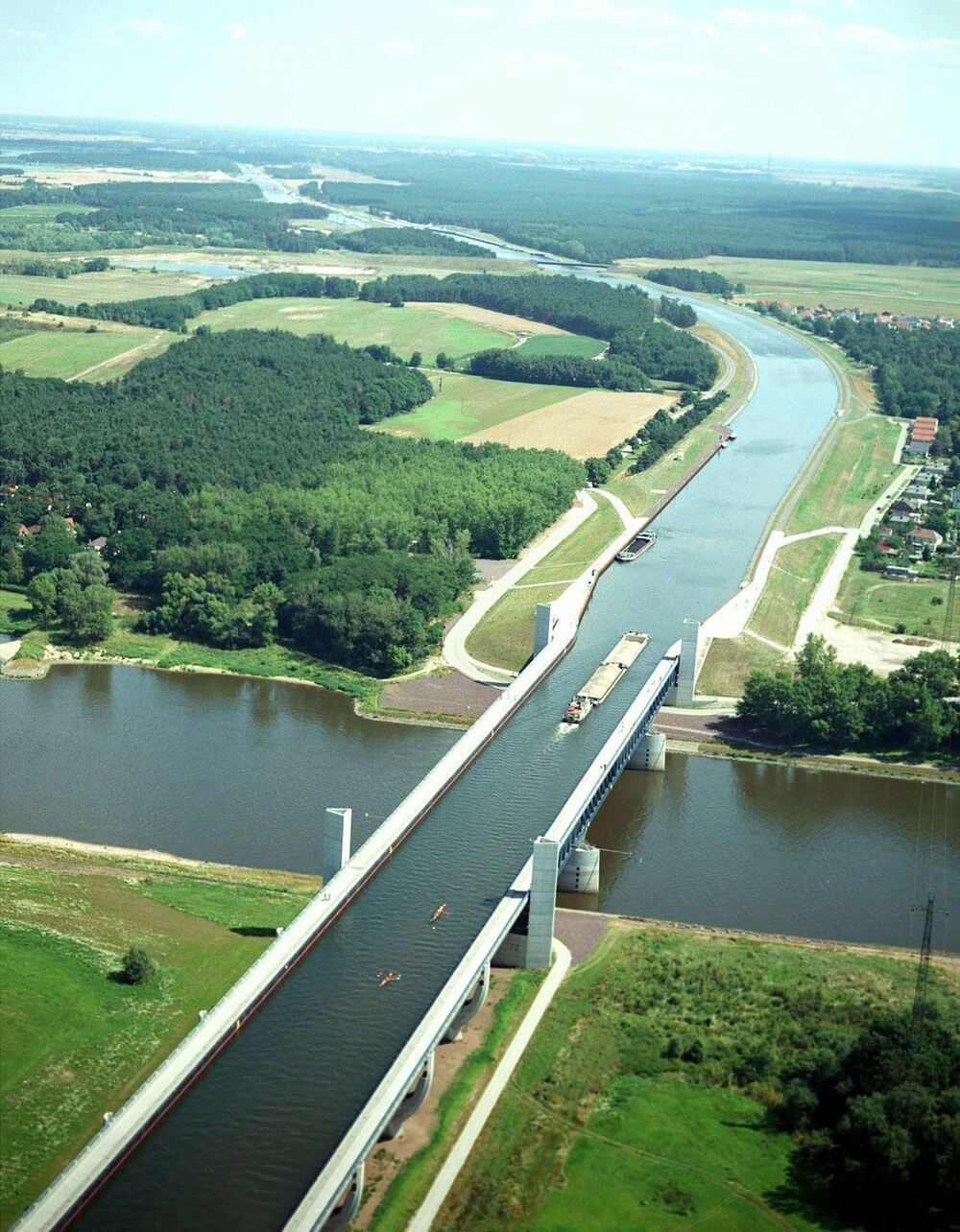 Aerial view of a water bridge carrying a canal over a river, with boats on the canal and green fields, trees, and buildings surrounding the area.