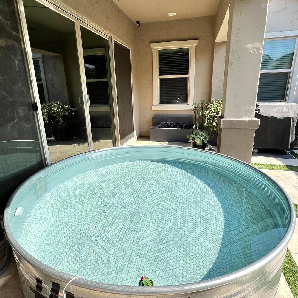 A large metal stock tank filled with clear water sits in a sunny, partially shaded patio area surrounded by house walls, windows, plants, and outdoor furniture.