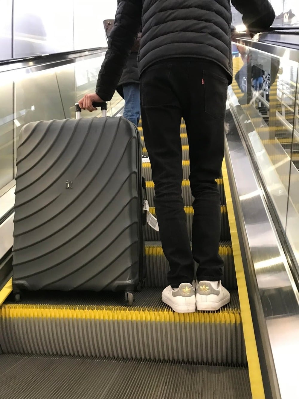 Person in black pants and white sneakers standing on an escalator, holding a large gray suitcase with a wavy pattern. The scene appears to be in an airport or travel hub.