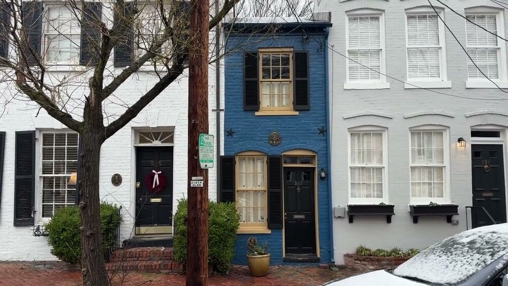 A narrow blue townhouse with black shutters is sandwiched between two white townhouses. A leafless tree and a wooden utility pole stand in front. A parked white car is partially visible on the right.