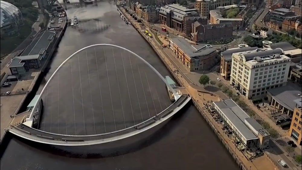 Aerial view of the Gateshead Millennium Bridge, tilted, spanning a river with nearby modern buildings and walkways on the riverbanks.