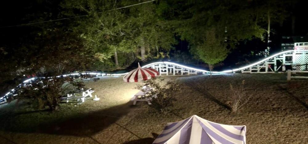 A nighttime scene shows a small roller coaster, lit by a string of lights, winding through a grassy area with picnic tables and tents. Trees are in the background.