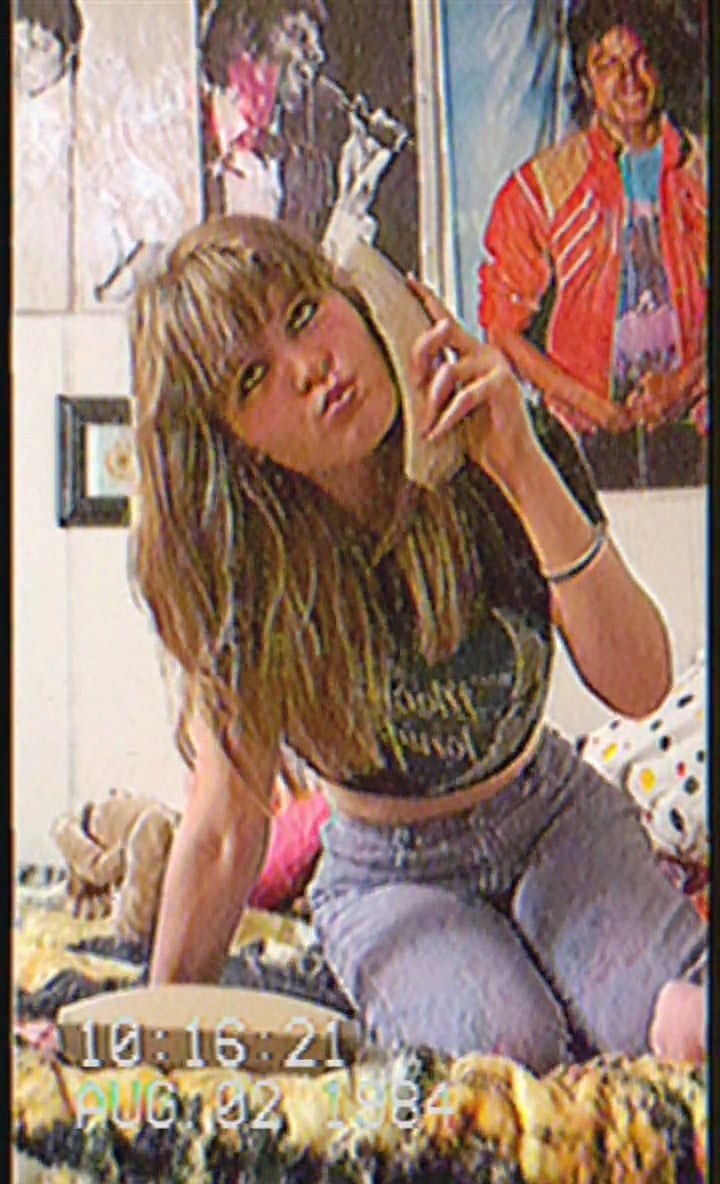 A young woman with long hair poses on a bed, holding a corded phone. She wears a crop top and jeans. Posters, including one of Michael Jackson, are on the wall. The timestamp reads “AUG 92 1984.”