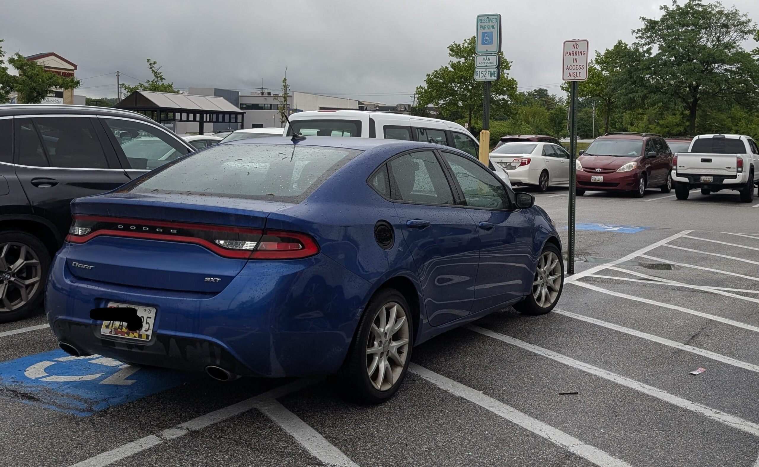 A blue Dodge sedan is parked over the line in a handicap parking space on a cloudy day. Other cars and handicap signs are visible in the parking lot.