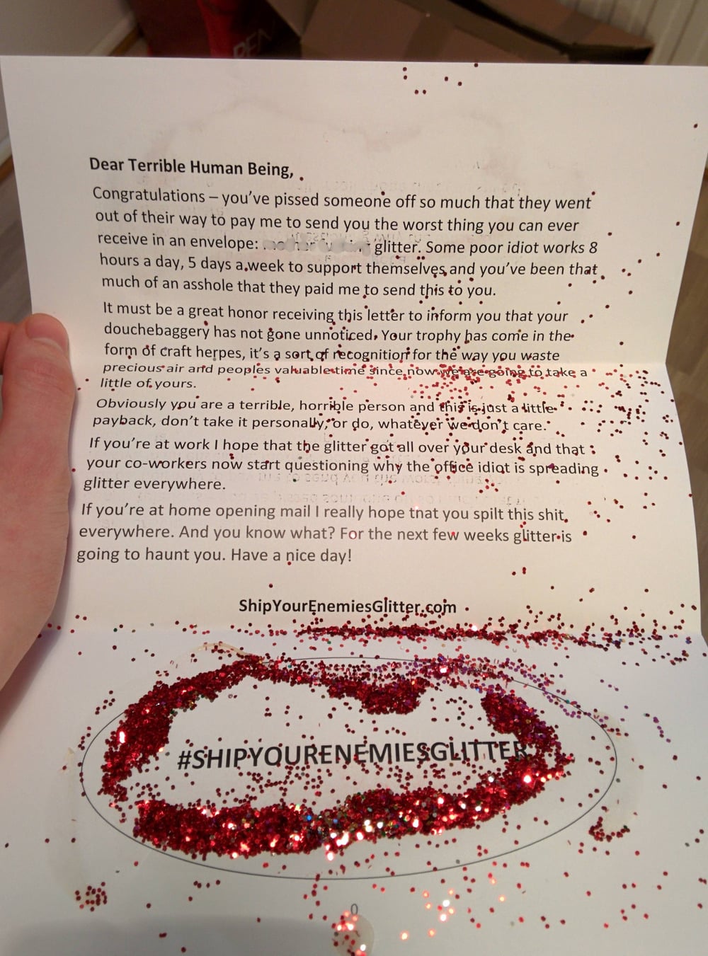 A hand holds a letter sprinkled with red glitter. The letter has a sarcastic message informing the recipient about glitter being sent to them, with “#SHIPYOURENEMIESGLITTER” printed at the bottom, surrounded by loose glitter.