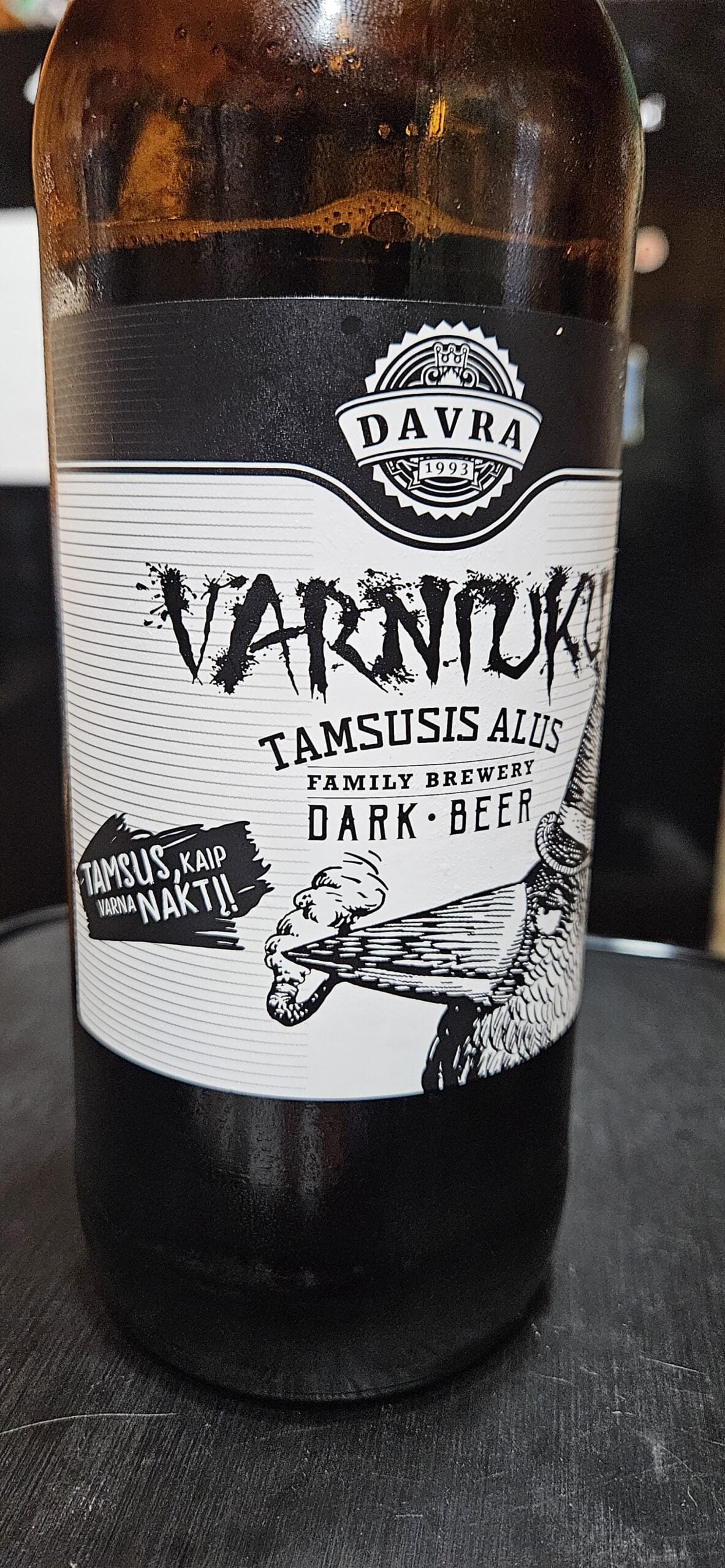 A close-up of a Davra Varniku Tamsusis Alus dark beer bottle, featuring a monochrome label with stylized text and an illustration of a hand pointing to the left.