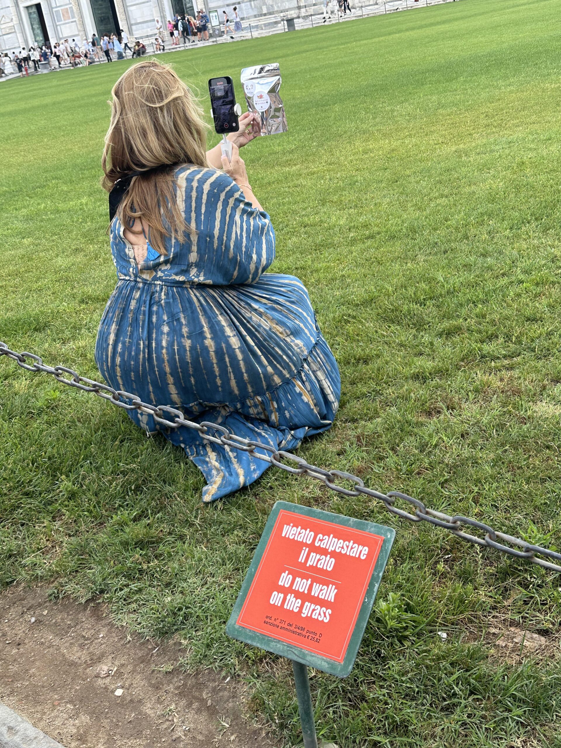 A woman in a blue dress kneels on grass to take a photo, near a red sign that says “Do not walk on the grass” in English and Italian.