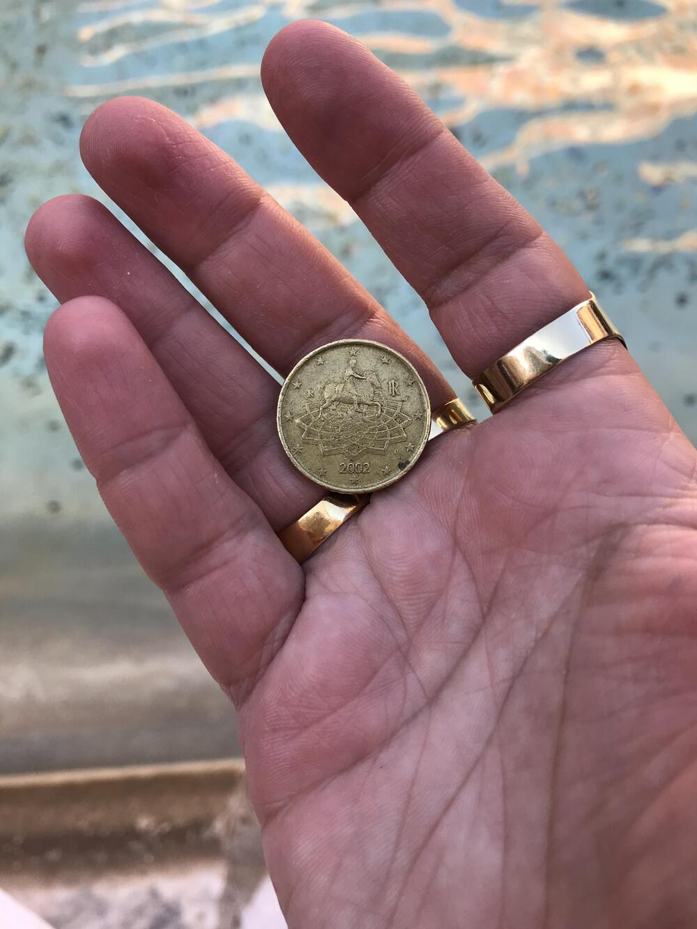 A hand with three gold rings holds a gold coin dated 2022, with a blurred background of water.