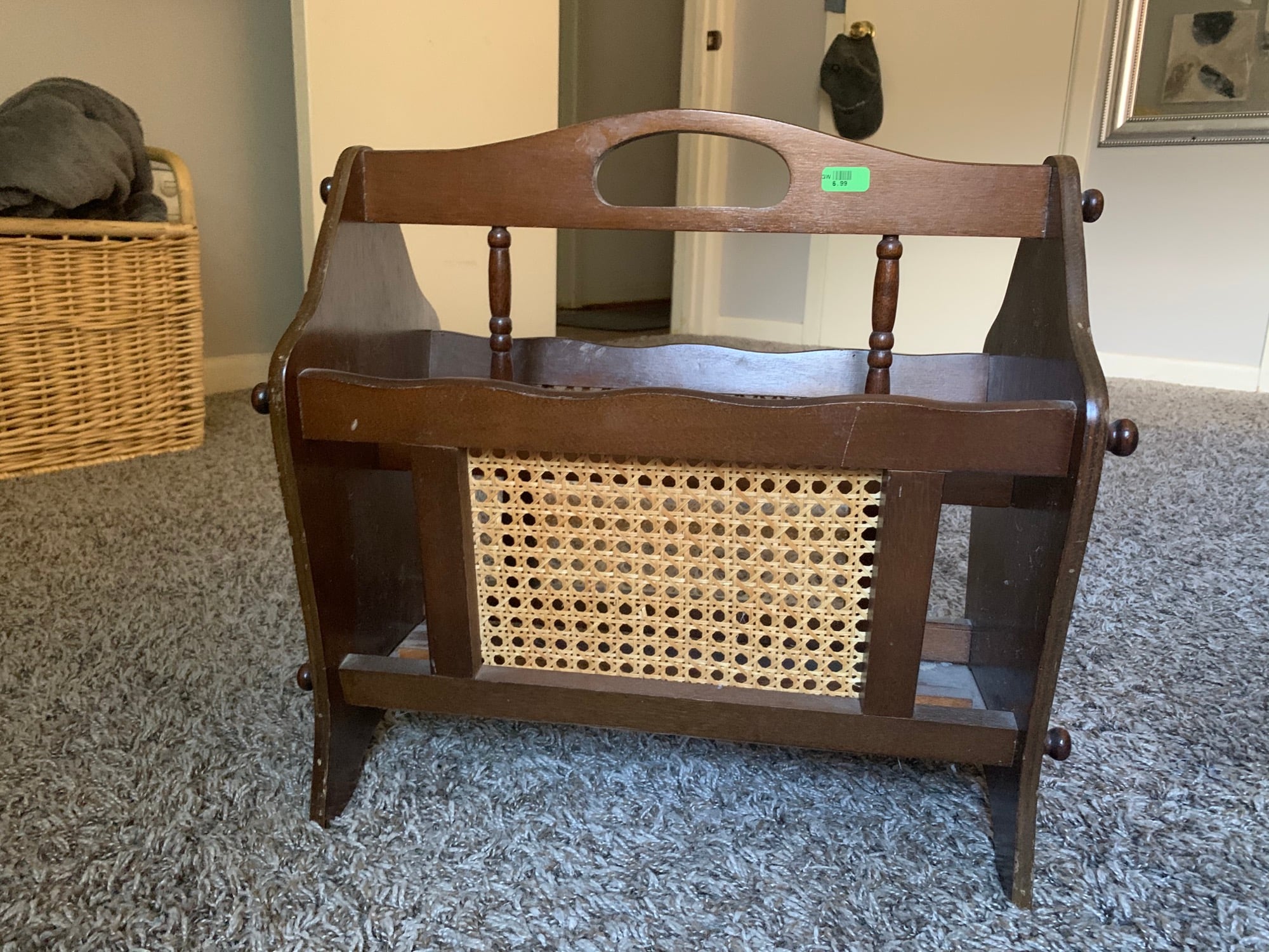 A wooden magazine rack with a woven cane panel sits on a carpeted floor in a room with a wicker basket, door, and mirror in the background.