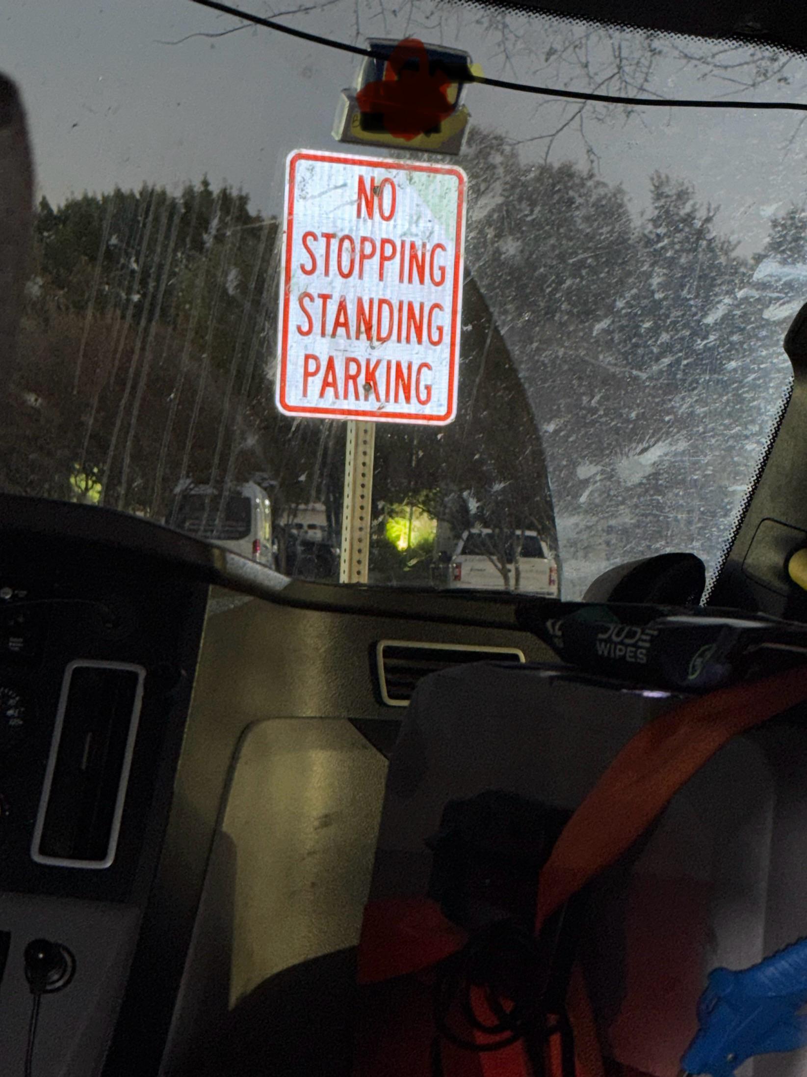 A red and white sign reads “No Stopping Standing Parking,” seen through a car windshield at night, with trees and parked cars in the background.