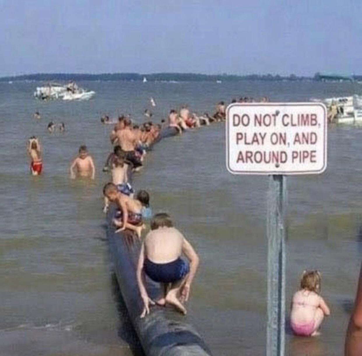 Children and adults climb and sit on a large pipe in the water, ignoring a nearby sign that says, “DO NOT CLIMB, PLAY ON, AND AROUND PIPE.” Boats are visible in the background.