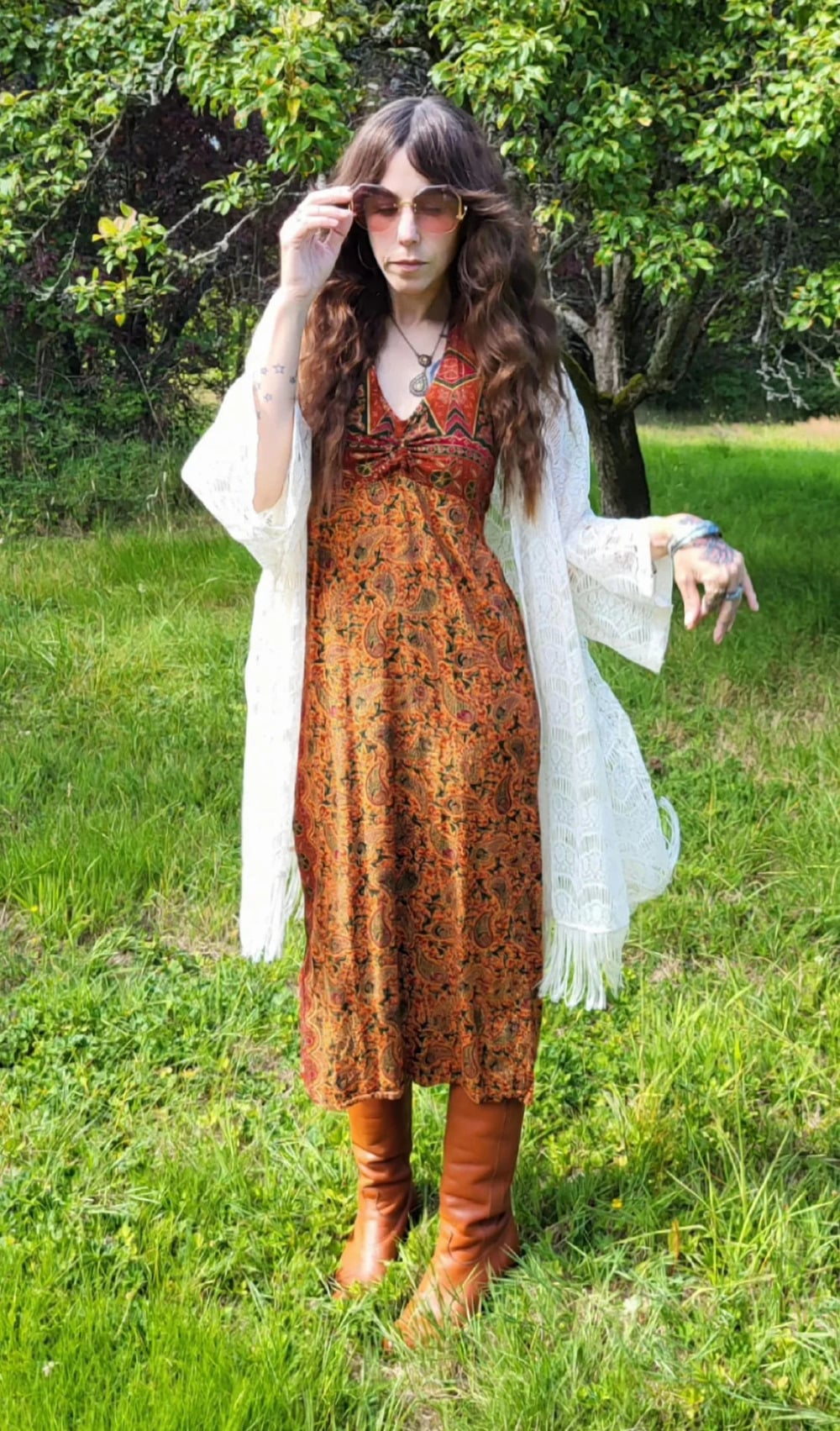 Person standing on grass, wearing a long patterned dress, brown boots, sunglasses, and a white shawl. Trees and greenery are in the background. The person is holding their sunglasses and looking at the camera.