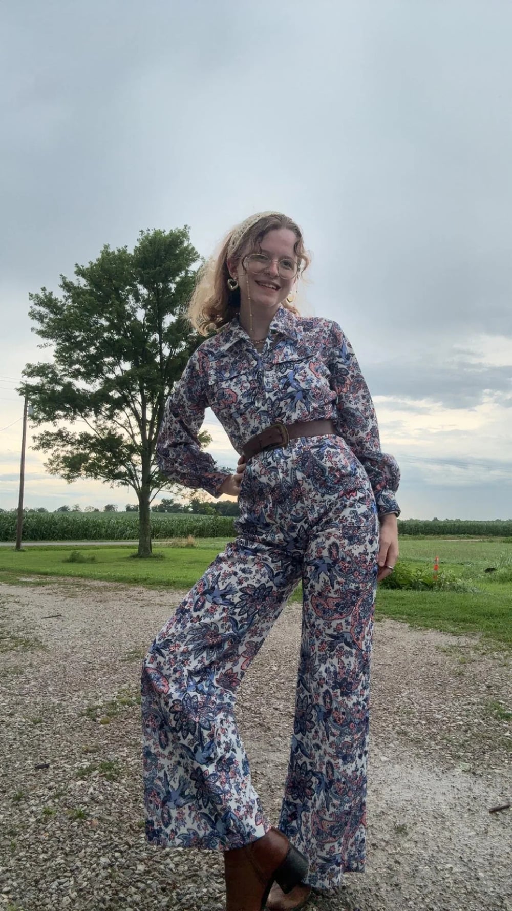 A person wearing a patterned jumpsuit and glasses stands on a gravel path, smiling with one hand on their hip. There’s a tree, green field, and cloudy sky in the background.