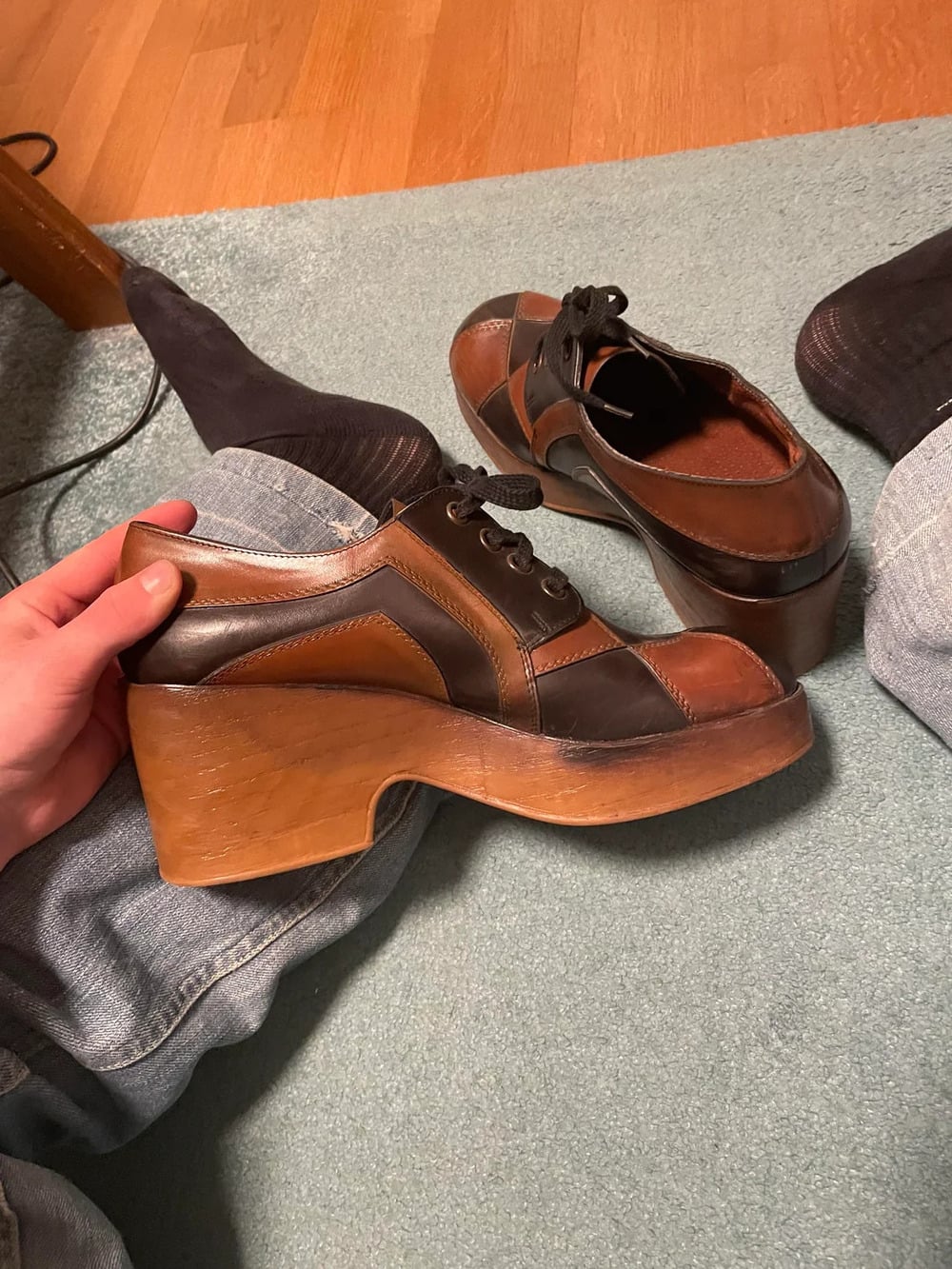 A person holds a brown and black platform shoe with a wooden heel. Another matching shoe rests on a blue carpet. The person’s legs in jeans and socks are visible.