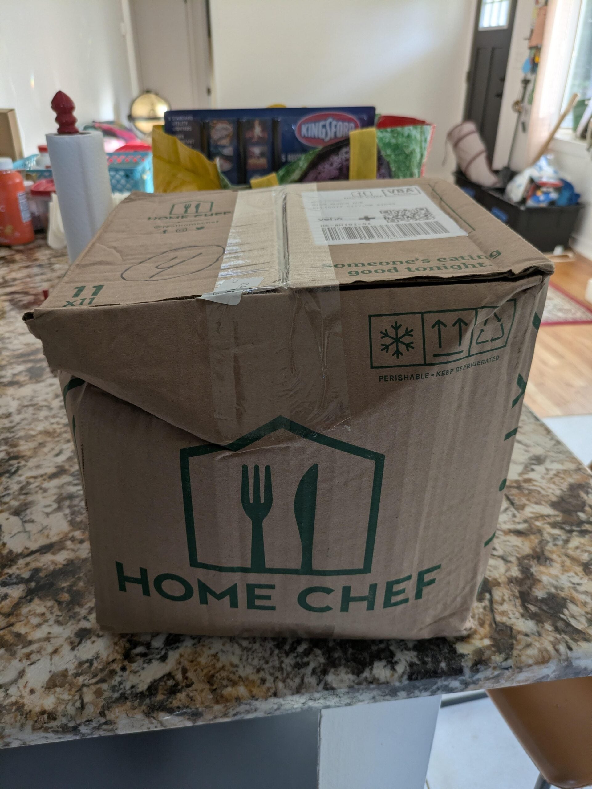 A damaged Home Chef delivery box sits on a kitchen counter. The box is dented and crumpled at the top, with visible shipping labels and branding. Various household items are in the background.