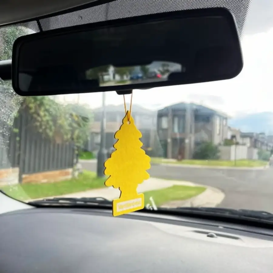 A yellow tree-shaped air freshener hangs from a car’s rearview mirror. Residential houses and a street are visible outside the windshield.