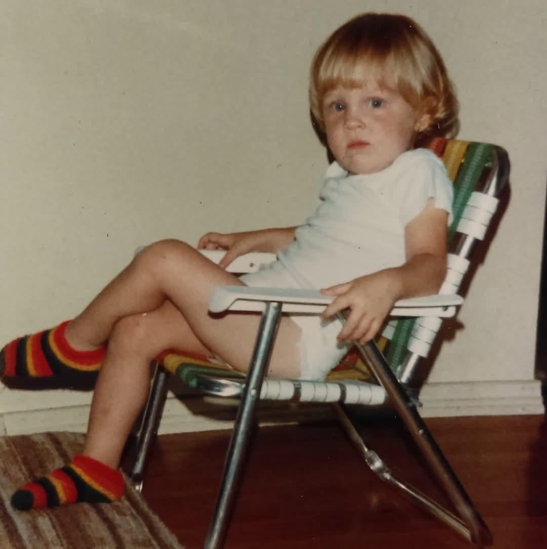 A young child with blond hair sits in a striped lawn chair indoors, wearing a white shirt, diaper, and colorful striped socks, looking at the camera with a serious expression.