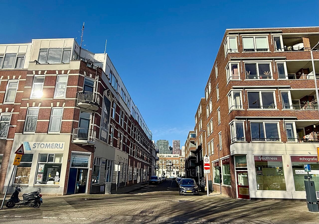 Sunny street scene with brick apartment buildings on both sides, shops on the ground floor, a motorcycle parked at the corner, and a clear blue sky overhead.