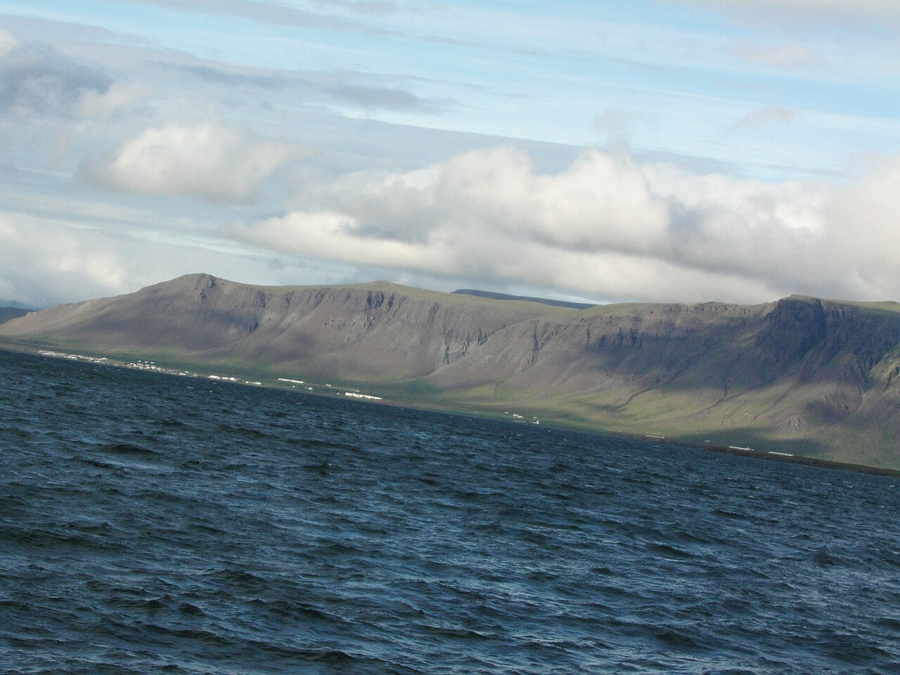 A choppy body of water in the foreground with distant mountains under a cloudy sky. The mountains appear rocky and partially covered in green vegetation.