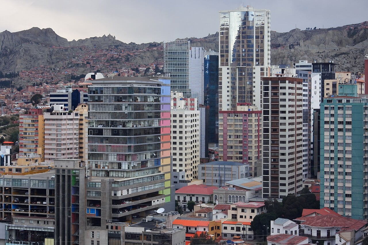 A cityscape with tall modern and older buildings closely packed together, set against a backdrop of hills under a cloudy sky.